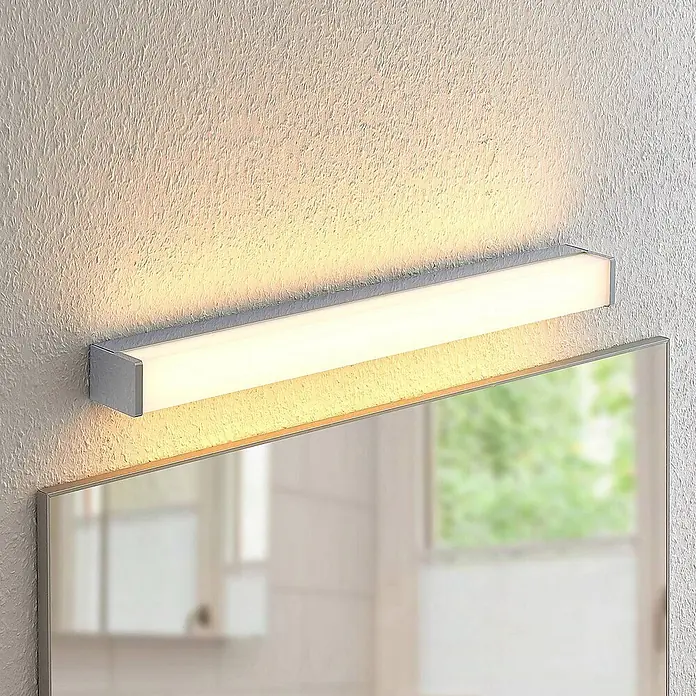 Lindby Wandleuchte Klea - L x B x H: 7 x 60 x 5 cm, chrom / silber - Eine moderne, rechteckige LED-Spiegelleuchte in Silberoptik, über einem Spiegel montiert, beleuchtet die strukturierte Wand und das Spiegelbild.