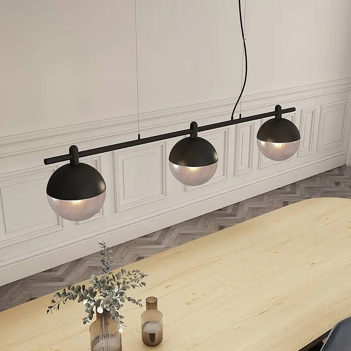Lucande Pendelleuchte Dustian - klar, Schwarz, E27 - Schwarze lineare Pendelleuchte mit drei kugelförmigen Lampen, halb schwarz, halb Klarglas, über einem hellen Holzesstisch.