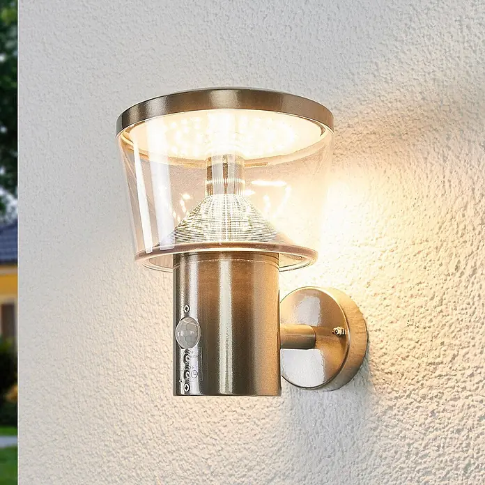 Lindby Außenwandleuchte Antje - alu / grau / zink, IP44 - Messingfarbene LED-Außenwandleuchte mit Bewegungsmelder, an rauer Wand befestigt.