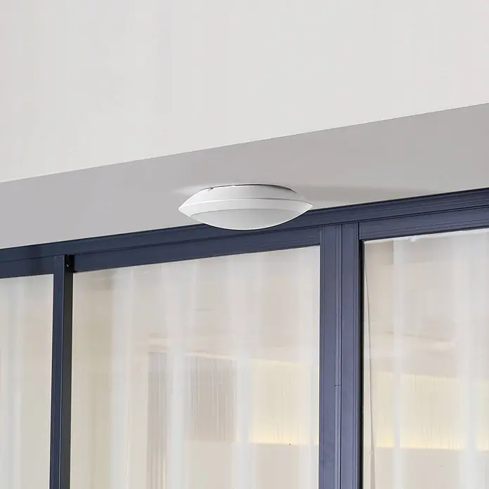 Lindby Außenwand- & Deckenleuchte Naira - weiß / opal, weiß / opal, IP65 - Weißes, flaches rundes LED-Deckenlicht über einer dunkel gerahmten Glaswand.