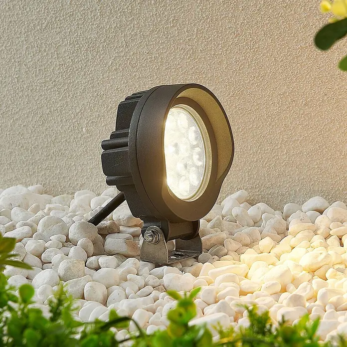 Lindby LED Wandleuchte innen in dunkelgrau, Wandstrahler -  - Schwarzer LED-Gartenstrahler mit sichtbaren weißen LEDs, beleuchtet weiße Kiesel und eine strukturierte Wand im Außenbereich.