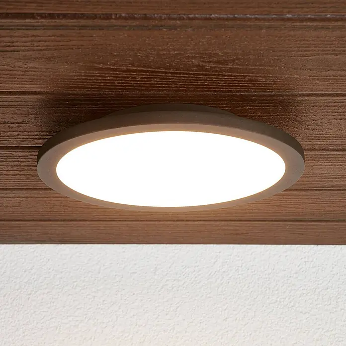 Lucande Außenwand- & Deckenleuchte Malena - schwarz, IP65 - Dunkelbronzene runde LED-Aufbauleuchte an Holzdecke, beleuchtet weiße Strukturwand.