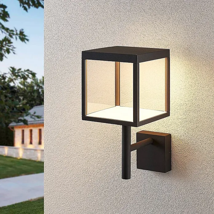 Lucande Außenwandleuchte Cube - schwarz, IP54 - Schwarze, würfelförmige Außenwandleuchte mit klaren Glaspaneelen, die warmes Licht auf eine strukturierte Wand wirft.