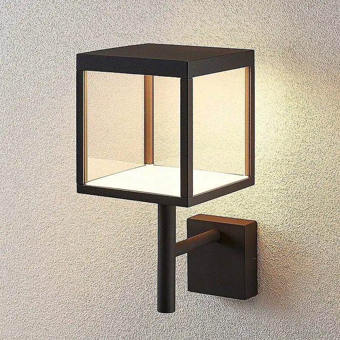 Lucande Außenwandleuchte Cube - schwarz, IP54 - Schwarze kubische Außenwandleuchte mit warmer Beleuchtung an einer strukturierten Wand montiert.