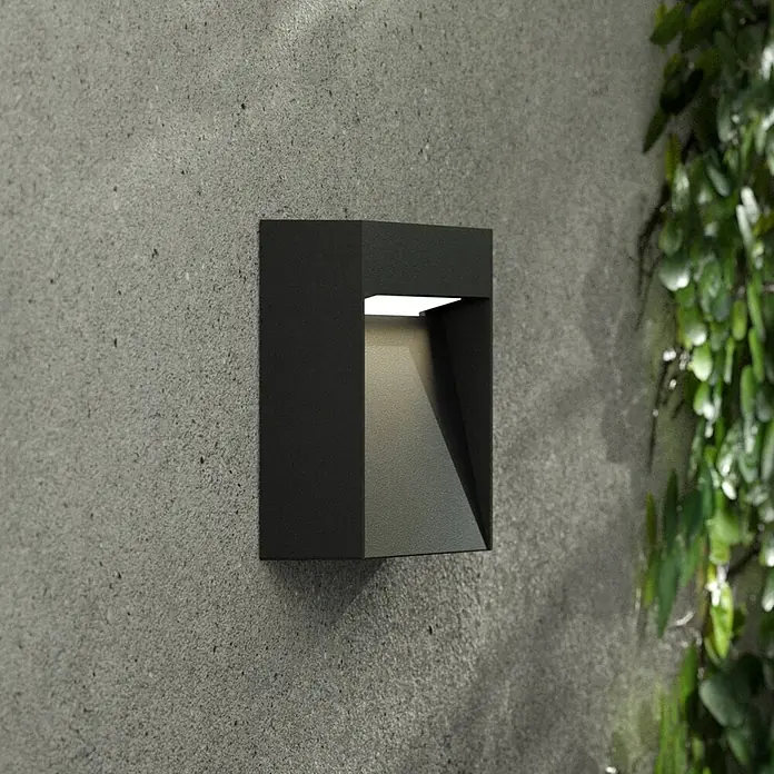 Lucande Außenwandleuchte Dijora - 3,1 x 9,5 x 9,5 cm, schwarz, IP65 - Schwarze moderne Wandleuchte an einer Betonwand, nach unten leuchtend, mit grünem Laub daneben.