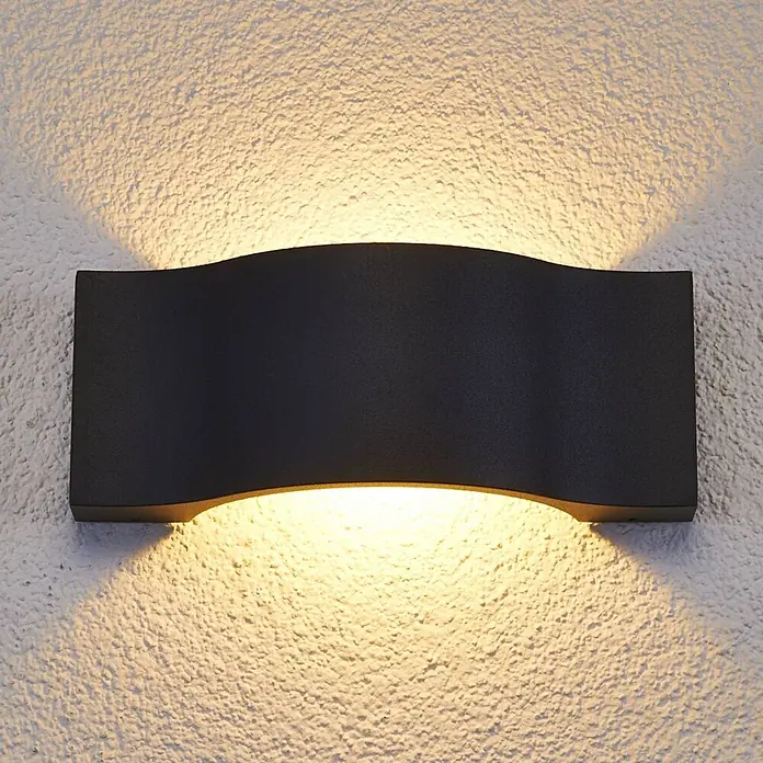 Lucande Außenwandleuchte Jace - schwarz, schwarz, IP54 - Schwarze wellenförmige Wandleuchte mit warmer Auf- und Abwärtsbeleuchtung an einer strukturierten Wand.