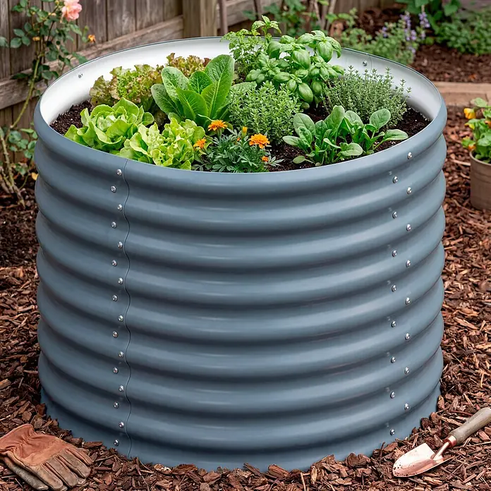 Vitavia Hochbeet Terra 630 - 82 x 82 x 63 cm, Zincalume®, Granit - Rundes, dunkelgraues Hochbeet aus Wellblech, gefüllt mit verschiedenen Grünpflanzen und kleinen orangefarbenen Blüten, in einem Garten mit Rindenmulch.