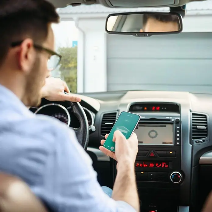 Hama Smartphone-Garagentoröffner 00176680 WLAN mit Sensor Steuerung per App/Sprachbefehl - Schwarz - Mann im Auto nutzt Smartphone mit grünem Display und Smart-Home-App zur Garagentorsteuerung.