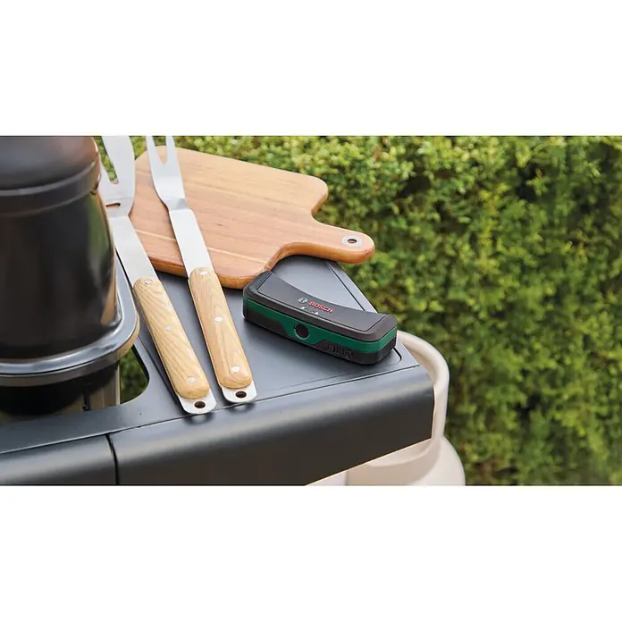 Bosch Gasflaschen-Füllstandsanzeiger Filio - Passend für: Stahl- und Aluminium-Flüssiggasflaschen (Propan und Butan) mit einem Durchmesser von 200-350 mm - Schwarzes und grünes Bosch Filio Gerät auf einem Tisch neben Grillgabeln und einem Holzschneidebrett.