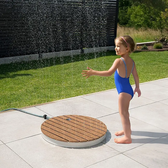 Home Deluxe Bodendusche CALIA - Teak - Runde Außendusche mit Holzlatten, sprüht Wasser, während ein Kind spielt. An Gartenschlauch angeschlossen.