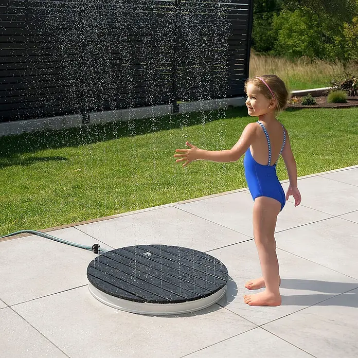 Home Deluxe Bodendusche CALIA - Anthrazit - Runder, schwarzer Latten-Gartenduschmatte, die Wasser sprüht und von einem Kind genutzt wird.