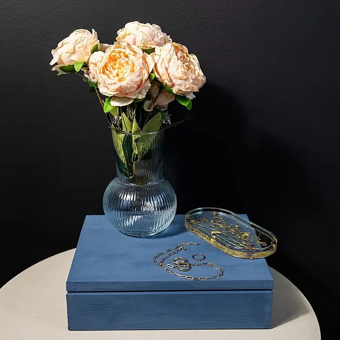 Creative Deco Speziallack Kreidefarbe für Möbel Jeans Blau - Blau, Matt, 1 l, Wasserbasiert - Eine blaue Holzschachtel dient als Basis für eine Glasvase mit pfirsichfarbenen Blumen, Goldschmuck und einem Harztablett mit Trockenblumen auf einem weißen Tisch.