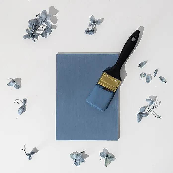 Creative Deco Speziallack Kreidefarbe für Möbel Jeans Blau - Blau, Matt, 1 l, Wasserbasiert - Ein schwarzer Pinsel mit blauer Farbe liegt auf einem blau bemalten Holzblock, umgeben von getrockneten blauen Blütenblättern.