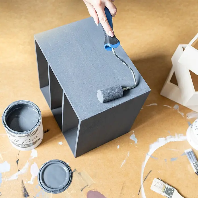 Creative Deco Speziallack Kreidefarbe für Möbel Anthrazitgrau - Grau, Matt, 1 l, Wasserbasiert - Eine Hand streicht einen Holz-Organizer mit Fächern grau mit einer Rolle. Eine offene Farbdose steht daneben.