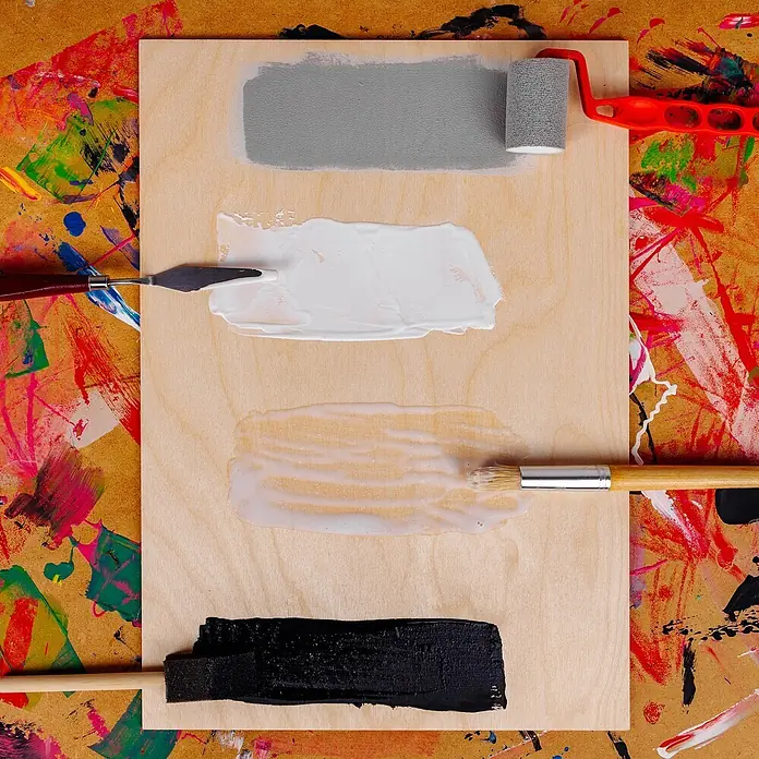 Creative Deco Speziallack Gesso Schwarz 1000ml - Schwarz, 1 l, Wasserbasiert - Helle Holzplatte mit grauen, weißen, hellbeigen und schwarzen Farbmustern, aufgetragen mit Rolle, Spachtel, Pinsel und Schwamm, auf einer farbgesprenkelten Oberfläche.
