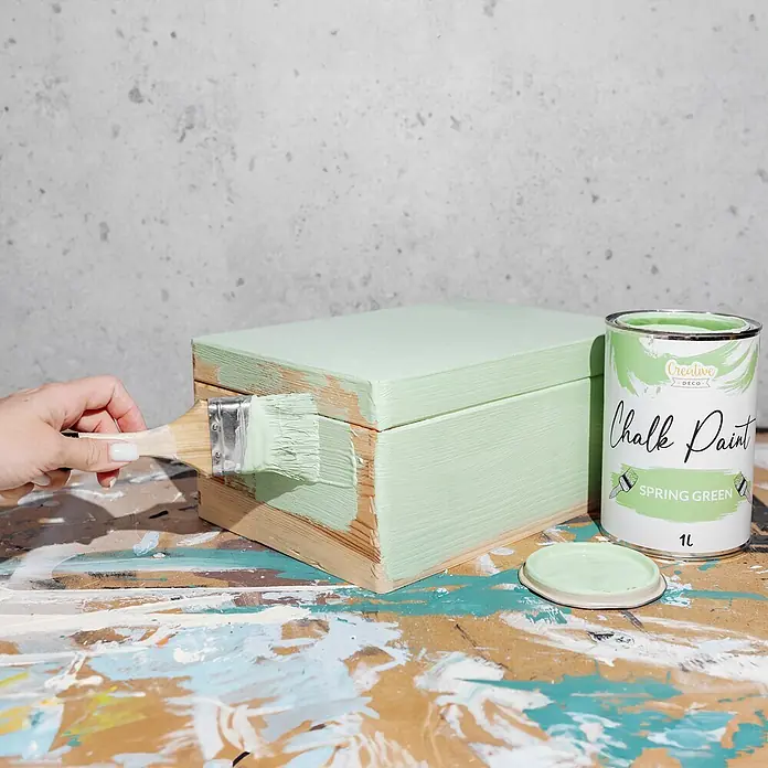 Creative Deco Speziallack Kreidefarbe für Möbel Frühlingsgrün - Grün, Matt, 1 l, Wasserbasiert - Eine Hand streicht eine Holzbox hellgrün mit einem Pinsel. Eine offene Dose "Spring Green" Kreidefarbe steht auf einem bemalten Tisch.