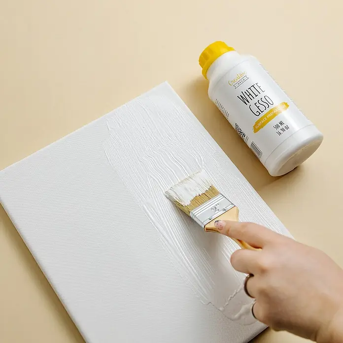 Creative Deco Speziallack Gesso Weiß 500ml - Weiß, 0,5 l, Wasserbasiert - Eine Hand bürstet weiße Gesso auf eine Leinwand, neben einer Flasche weißer Gesso.