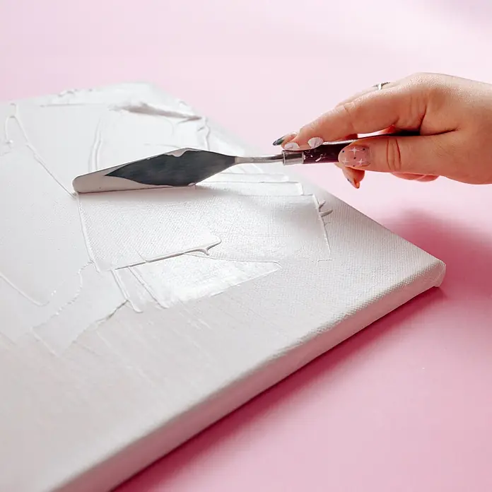Creative Deco Speziallack Gesso Klar 500ml - Transparent, 0,5 l, Wasserbasiert - Eine Hand verwendet ein Spachtelmesser, um weiße Modellierpaste auf eine weiße Leinwand auf einem rosa Hintergrund zu streichen.