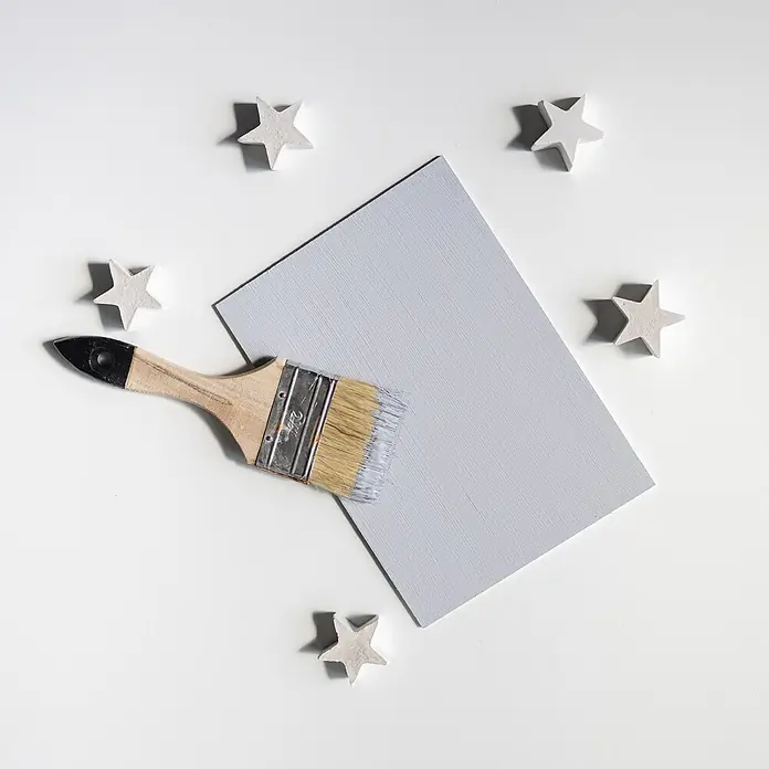 Creative Deco Speziallack Kreidefarbe für Möbel Grau - Grau, Matt, 1 l, Wasserbasiert - Pinsel mit hellgrauer Farbe auf einer bemalten Probe, umgeben von sternförmigen Dekorationen.