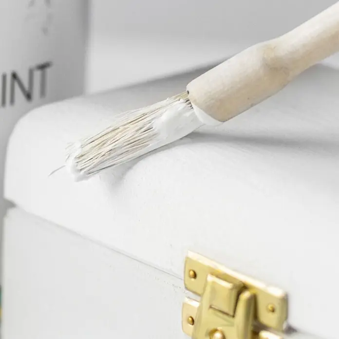 Creative Deco Speziallack Kreidefarbe für Möbel Weiß - Weiß, Matt, 1 l, Wasserbasiert - Pinsel trägt weiße Farbe auf eine weiße Holzoberfläche auf, mit einem goldenen Verschluss.