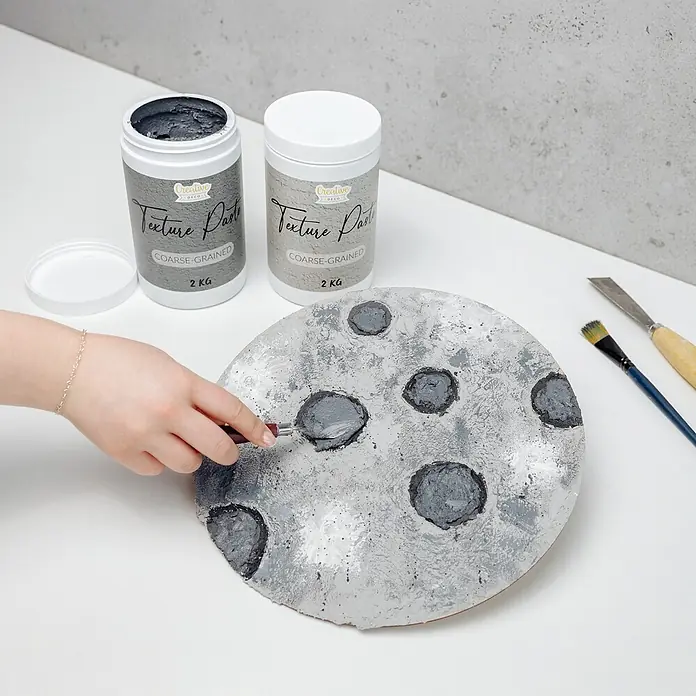 Creative Deco Speziallack Strukturpaste Grobkörnig 2 kg Grau - Grau, Sonstiges, 2 l, Sonstiges - Eine Hand trägt grobkörnige Texturpaste von Creative Deco auf eine runde, mondähnliche Fläche auf. Zwei 2KG-Dosen sind sichtbar.