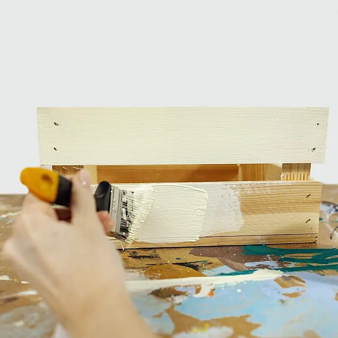 Creative Deco Speziallack Kreidefarbe für Möbel Vanillegelb - Gelb, Matt, 1 l, Wasserbasiert - Eine Hand streicht eine Holzkiste mit einem Pinsel cremeweiß. Die Kiste ist teilweise bemalt.