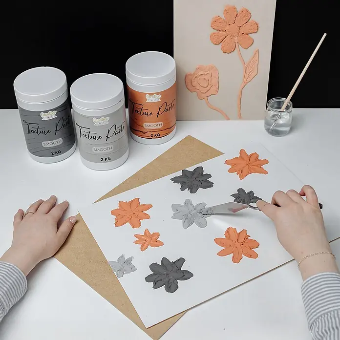 Creative Deco Speziallack Strukturpaste Glatt 2 kg Ziegelrot - Rot, Sonstiges, 2 l, Sonstiges - Hände tragen graue und orange Strukturpaste aus Behältern auf, um Blumenkunst zu schaffen.