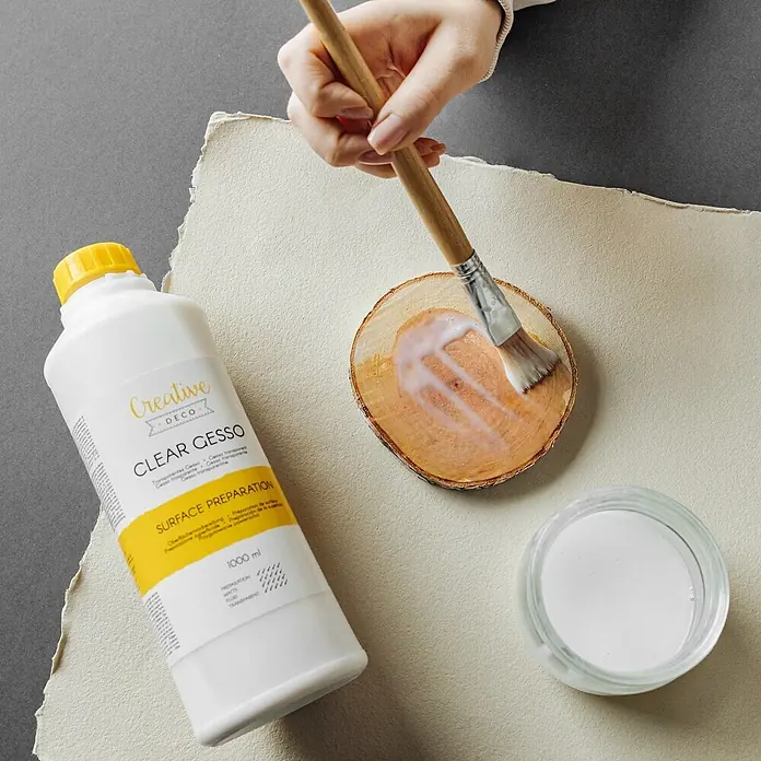 Creative Deco Speziallack Gesso Klar 1000ml - Transparent, 1 l, Wasserbasiert - Hand bürstet klares Gesso aus einer Flasche auf eine Holzscheibe. Ein Glas Gesso ist in der Nähe.