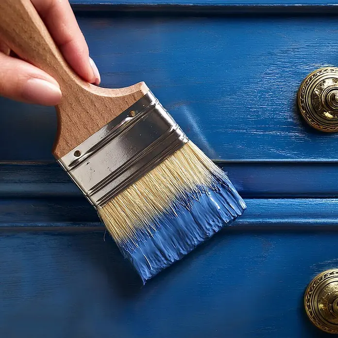 Creative Deco Speziallack Kreidefarbe für Möbel Ozeanblau - Blau, Matt, 1 l, Wasserbasiert - Eine Hand streicht mit einem Pinsel blaue Farbe auf eine blaue Holzkommode mit goldenem Knopf.