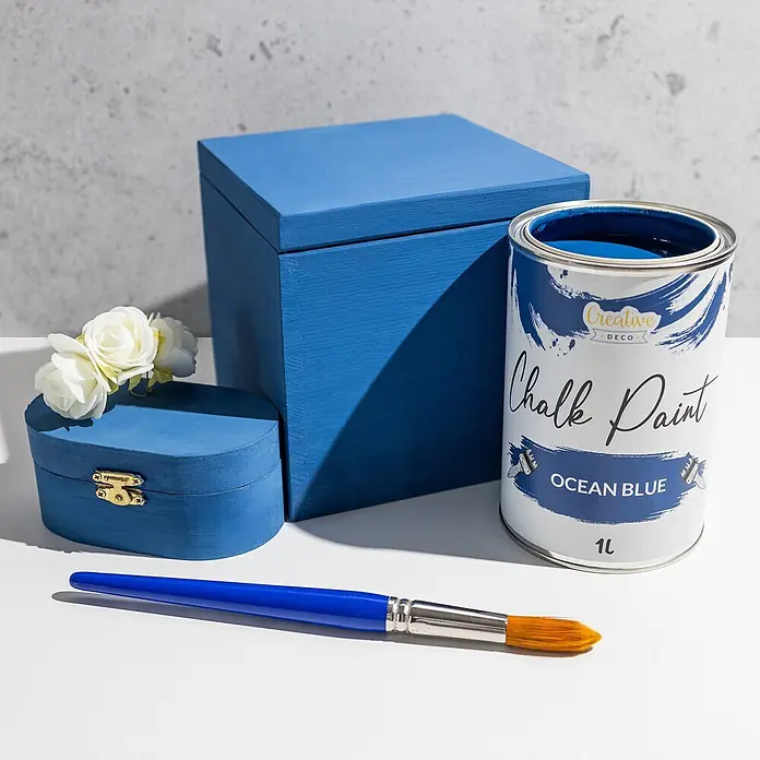 Creative Deco Speziallack Kreidefarbe für Möbel Ozeanblau - Blau, Matt, 1 l, Wasserbasiert - Ozeanblaues Kreidefarbendose, zwei blaue Holzboxen (eine oval, eine rechteckig), ein blauer Pinsel und weiße Blumen.