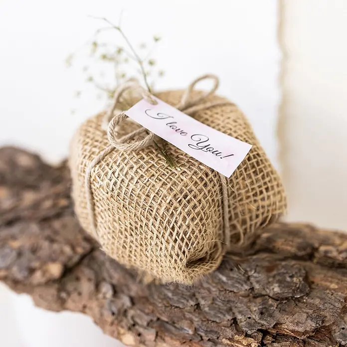 Creative Deco Bastelband Juteband 20cm x 10m - Braun - Kleines Geschenk in natürlichem Jute-Stoff, mit Schnur gebunden, rosa 'I love You!'-Anhänger und Schleierkraut, auf Rinde liegend.