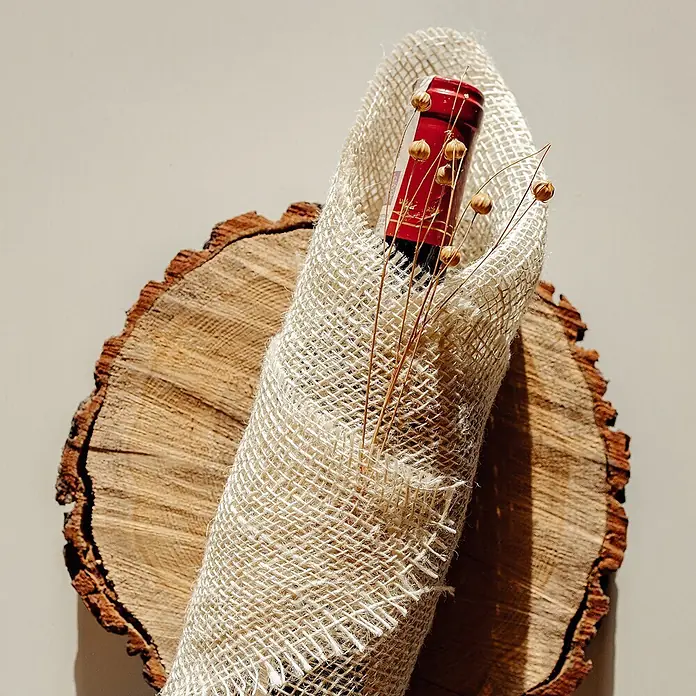 Creative Deco Bastelband Juteband Weiß 20cm x 10m - Weiß, 10 m - Rote Flasche in naturfarbenem Gewebe mit getrockneten Pflanzenakzenten auf einer runden Holzscheibe.