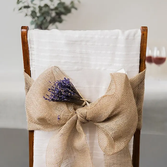 Creative Deco Bastelband Juteband 20cm x 10m - Braun - Ein Holzstuhl mit weißem Bezug, geschmückt mit einer großen Schleife aus Sackleinen und einem Bündel getrockneten Lavendels.