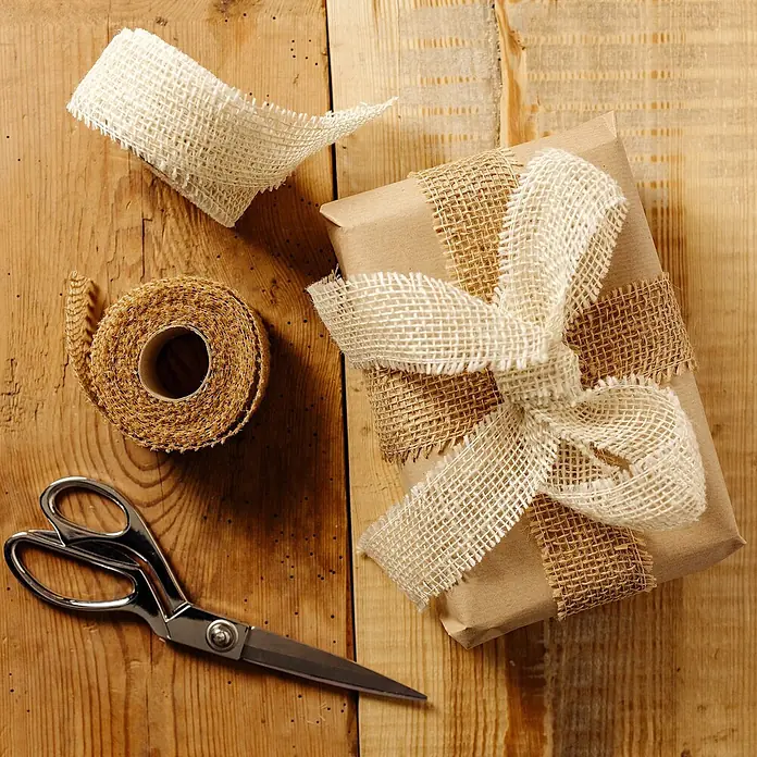 Creative Deco Bastelband Juteband Weiß 2 Rollen 5cm x 5m - Weiß, 10 m - Braun eingepacktes Geschenk mit beigefarbenen Sackleinenbändern und Schleife, dazu Sackleinenrollen und Silberschere auf rustikaler Holzoberfläche.