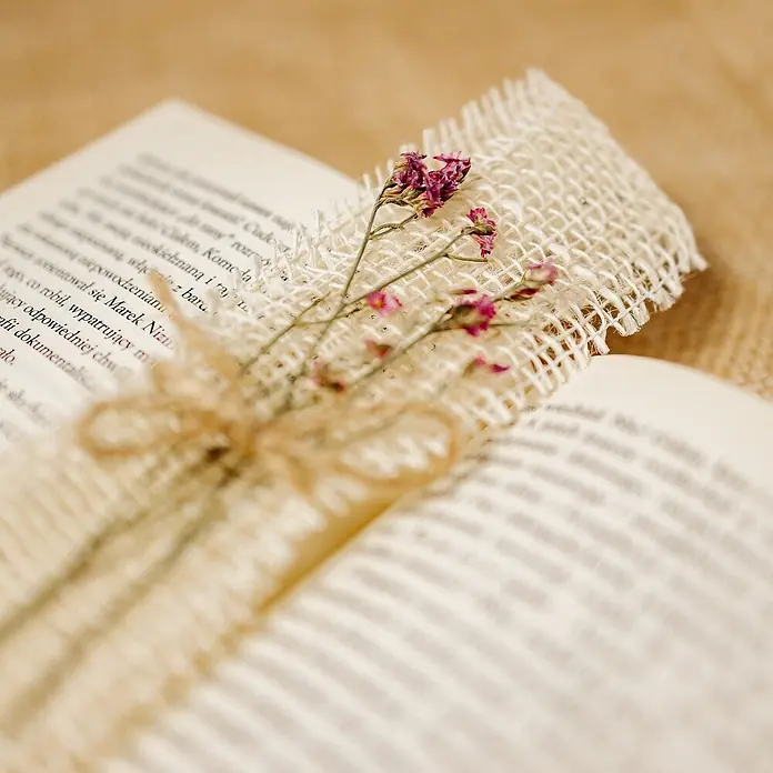 Creative Deco Bastelband Juteband Weiß 2 Rollen 5cm x 5m - Weiß, 10 m - Ein rustikales Lesezeichen aus gewebtem Sackleinen und getrockneten rosa Blüten in einem offenen Buch.