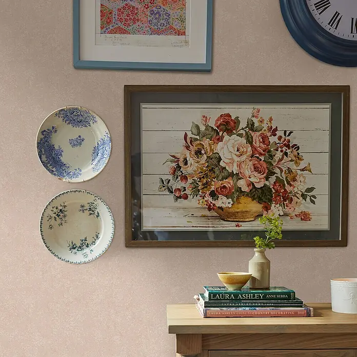 Laura Ashley Vliestapete Vliestapete Brindley - Rosa, Uni, 10 x 0,52 m - Eine hellrosa, strukturierte Tapete auf einer Wand mit gerahmter Kunst, Ziertellern und einer blauen Uhr, mit einem Holztisch.