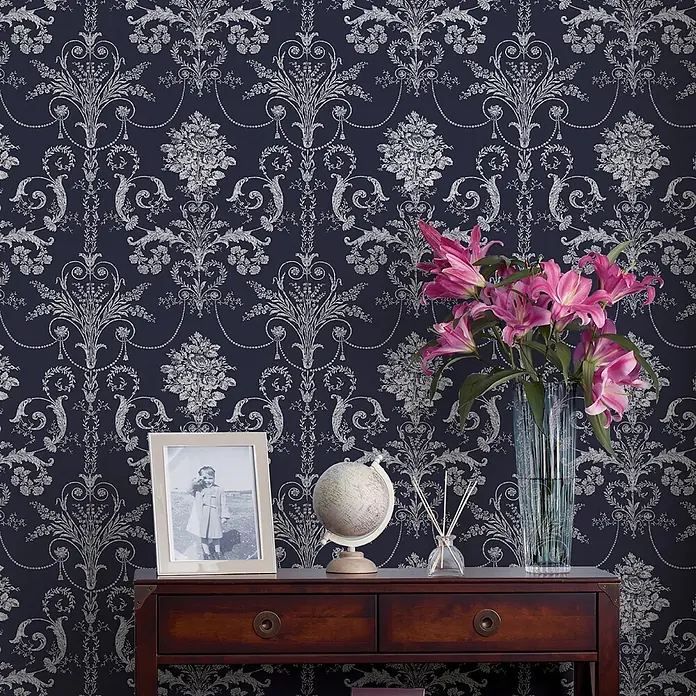 Laura Ashley Vliestapete Vliestapete Josette Damast - Dunkelblau, Ornament, 10 x 0,52 m - Dunkelblaue Tapete mit aufwendigem silbernem Damastmuster, an einer Wand hinter einem Holztisch mit Dekoration.