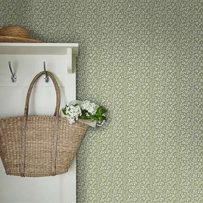 Laura Ashley Vliestapete Vliestapete Süßes Alyssum - Grün, Floral - Grüne Tapete mit weißem Blumenmuster an einer Wand, neben einem weißen Garderobenhaken mit Korbtasche und Hut.