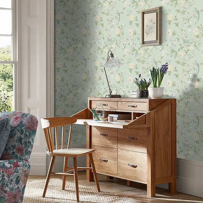 Laura Ashley Vliestapete Vliestapete Sommerpalast - Grün, Floral - Helles Eichensekretär mit ausklappbarer Schreibfläche und mehreren Schubladen, kombiniert mit einem passenden Holzstuhl in einem Home-Office-Ambiente.