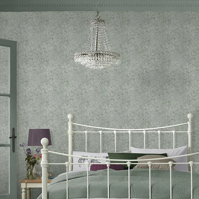 Laura Ashley Vliestapete Corrina Blatter - Grün, Floral, 1.000 x 52 cm - Grüne Tapete mit subtilem Blattmuster, verwendet in einem Schlafzimmer mit weißem Metallbett und Kronleuchter.