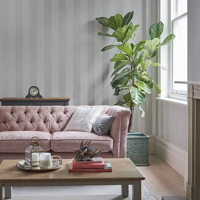 Laura Ashley Vliestapete Vliestapete Lille - Silber, Ornament - Ein gemütliches Wohnzimmer mit einem rosa getufteten Sofa, grau gestreifter Tapete und einer großen Pflanze in einem Ziertopf.