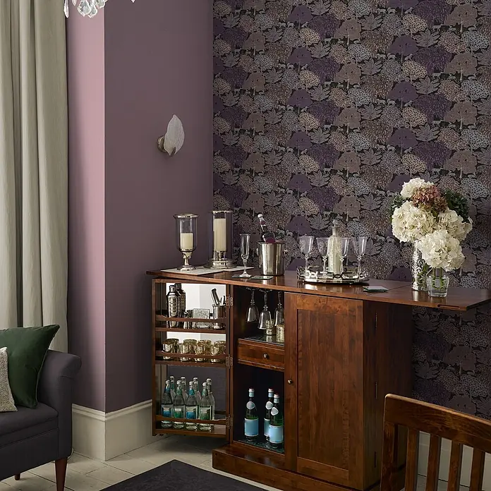Laura Ashley Vliestapete Vliestapete Garwood Grove - Lila, Floral, 10 x 0,52 m - Dunkelbrauner Holz-Barschrank, geöffnet mit Flaschen, Gläsern und Barzubehör in einem Raum.
