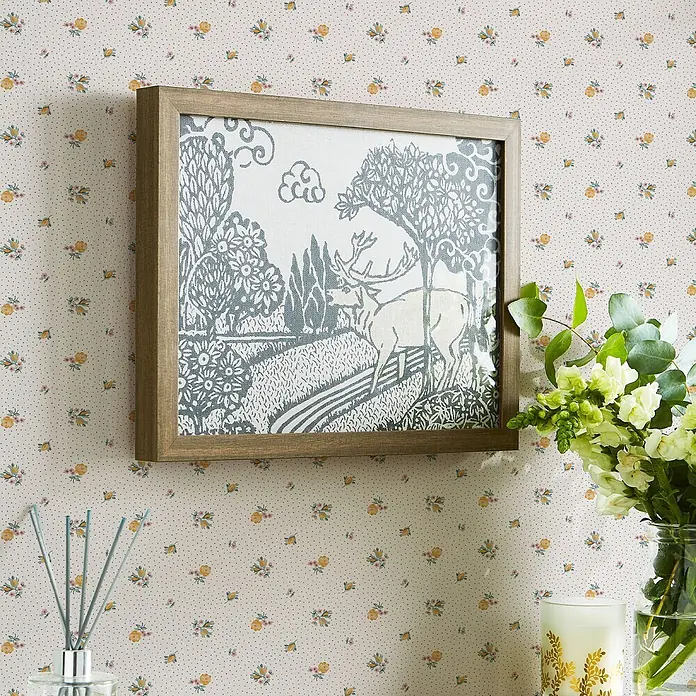 Laura Ashley Vliestapete Claudias Freund - Gelb, Floral, 1.000 x 52 cm - Ein gerahmtes Bild mit Holzrahmen und einer blau-weißen Hirschillustration in einer Landschaft, an einer gemusterten Wand hängend.