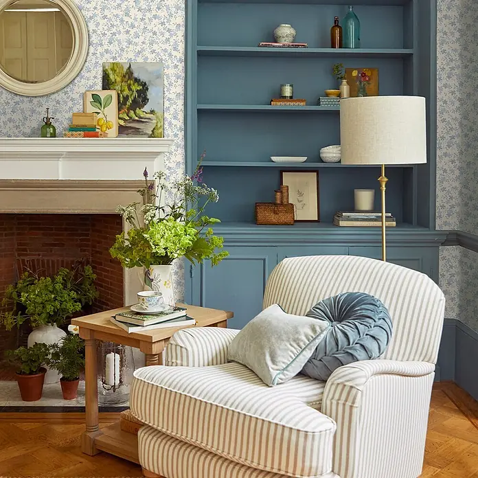 Laura Ashley Vliestapete Misterton Trail - Hellblau, Floral, 1.000 x 52 cm - Gemütlicher Sessel mit cremefarbenen und hellbraunen Streifen, mit hellgrauem und plissiertem blauem Kissen, in einem stilvollen Wohnzimmer.