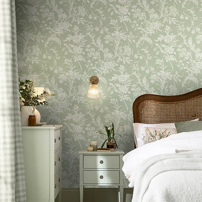 Laura Ashley Vliestapete Vliestapete gewebtes Blumen - Grün, Floral, 10 x 0,52 m - Hellgrüne Tapete mit weißem Blumen- und Rankenmuster schmückt eine Schlafzimmerwand hinter Bett und Nachttischen.