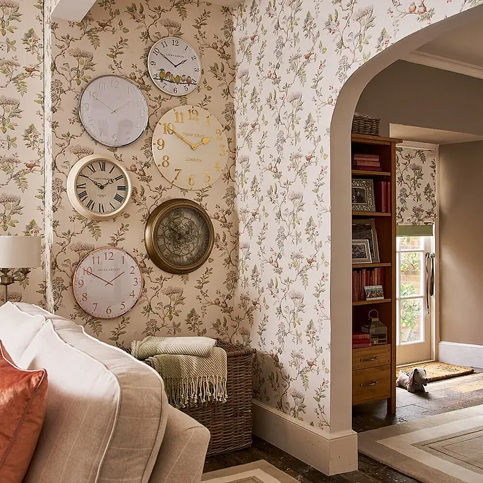Laura Ashley Vliestapete Vliestapete Elderwood - Mehrfarbig, Floral - Eine Sammlung dekorativer Wanduhren in verschiedenen Stilen und Größen schmückt eine gemusterte Wand in einem Wohnzimmer und verleiht dem Dekor Charme.