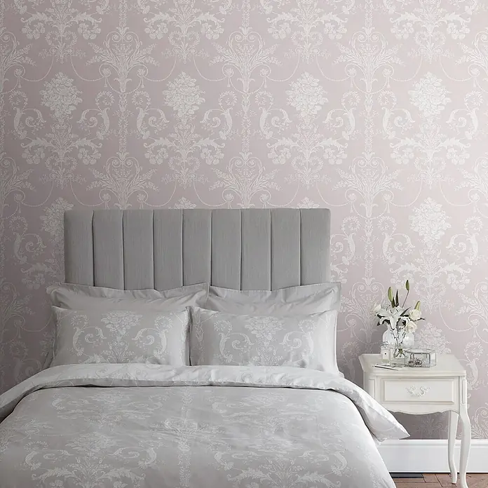 Laura Ashley Vliestapete Vliestapete Josette Damast - Amethyst/Rosa, Ornament - Graue Bettwäsche mit Damastmuster auf einem Bett mit grauem Polsterkopfteil, in einem Schlafzimmer mit rosa Damasttapete.