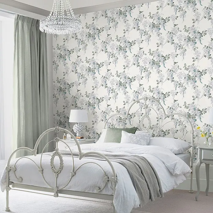 Laura Ashley Vliestapete Fliederzweige - Blau, Floral, 1.000 x 52 cm - Schlafzimmer mit heller floraler Tapete im Glyzinienmuster, ergänzt durch ein weißes Metallbett und hellgrüne Vorhänge.