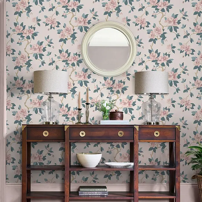 Laura Ashley Vliestapete orientalische Blüte - Leinen, Floral, 1.000 x 52 cm - Ein dunkler Holztisch mit zwei Lampen und einem runden Spiegel steht vor Blumentapete, darunter Regale mit Dekoration.