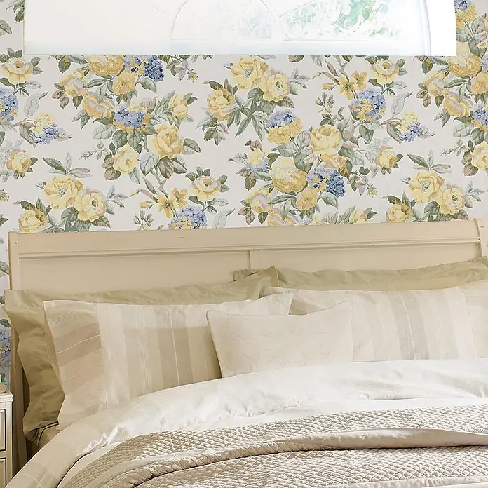 Laura Ashley Vliestapete Vliestapete Pembrey - Gold, Floral, 10 x 0,52 m - Ein Bett mit cremefarbenem Kopfteil, heller Bettwäsche und Kissen vor einer Blumenmuster-Tapete.
